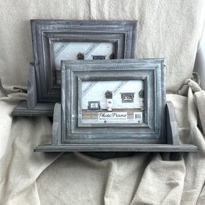 May include: Two grey wooden photo frames with a distressed finish. Each frame has a shelf and a back support. The frames hold 10 x 15 cm photos and are labelled "Table Top" and "Wall Mount".