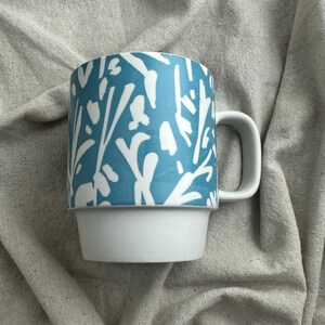 May include: A white and blue ceramic mug with a unique design. The top portion of the mug features a blue background with a white floral pattern. The bottom portion of the mug is white. The mug has a white handle.