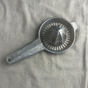 May include: A silver-colored citrus juicer with a long handle and a conical reamer. The juicer has a circular bowl with a series of vertical slots. The handle has a hole at the end for hanging. The juicer is made of metal.