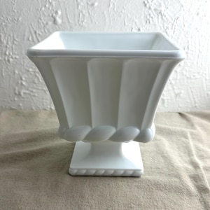 May include: White ceramic planter with a square top and a fluted design. The planter has a pedestal base with a rope-like design.