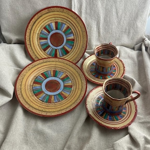 May include: A set of vintage ceramic tableware featuring two plates and two teacups with saucers. The plates and saucers have a yellow background with a red border and a colorful geometric design. The teacups have a matching design.