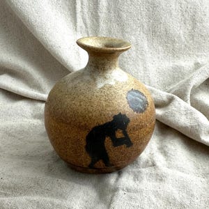 Peut inclure: Petit vase rond en céramique marron moucheté, avec un col étroit et un bord évasé. Le vase présente une silhouette noire d'une personne et un cercle gris foncé. Le vase a un aspect rustique et fait à la main.