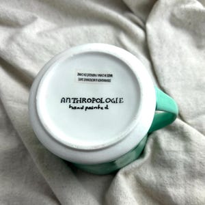 May include: A white ceramic mug with a turquoise handle and rim. The bottom of the mug features the text "ANTHROPOLOGIE hand painted" in black. The mug is labeled "DISHWASHER & MICROWAVE SAFE MADE IN CHINA/ FABRIQUE EN CHINE".