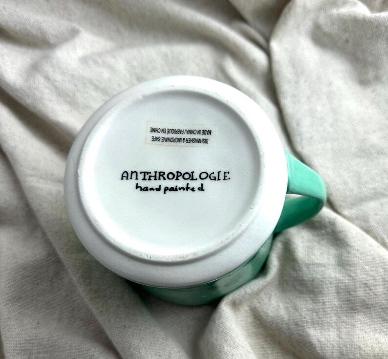 May include: A white ceramic mug with a turquoise handle and rim. The bottom of the mug features the text "ANTHROPOLOGIE hand painted" in black. The mug is labeled "DISHWASHER & MICROWAVE SAFE MADE IN CHINA/ FABRIQUE EN CHINE".