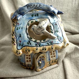 May include: Hand-painted ceramic birdhouse with a dove cutout and the word "peace" on top. The birdhouse features blue, yellow, and green floral designs. The roof has a scalloped edge, and the door has a decorative design.