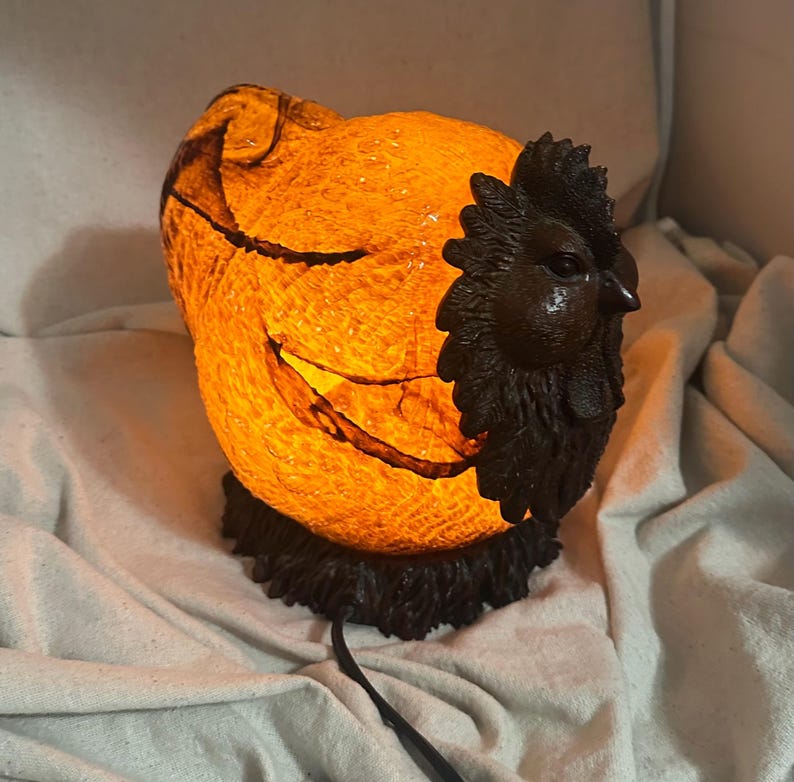 May include: A decorative lamp shaped like a rooster. The lamp has a textured, orange-colored body with a black rooster head and base. The lamp is lit, and the light shines through the body.
