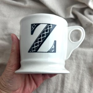 May include: White ceramic mug featuring a large, black letter "Z" with a decorative diamond pattern. The mug has a rounded base and a handle with a circular opening. A simple, elegant design.