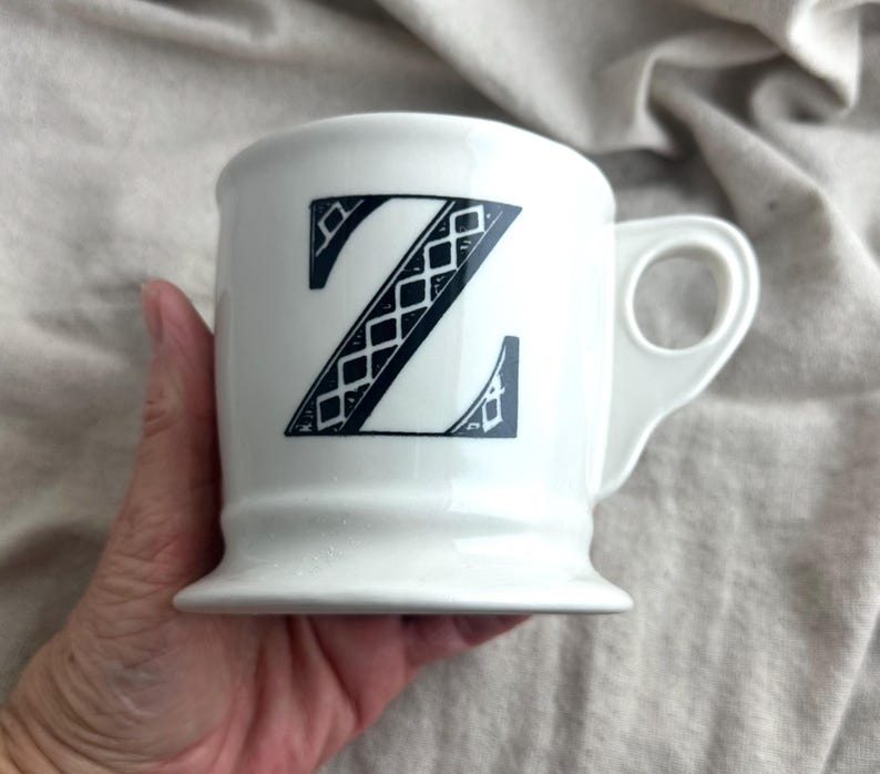 May include: White ceramic mug featuring a large, black letter "Z" with a decorative diamond pattern. The mug has a rounded base and a handle with a circular opening. A simple, elegant design.