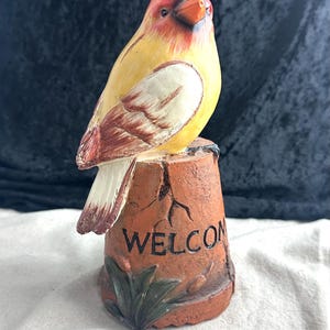 May include: A ceramic bird figurine with red, yellow, and white paint, perched on a brown pot with the word "WELCOME" painted on it.