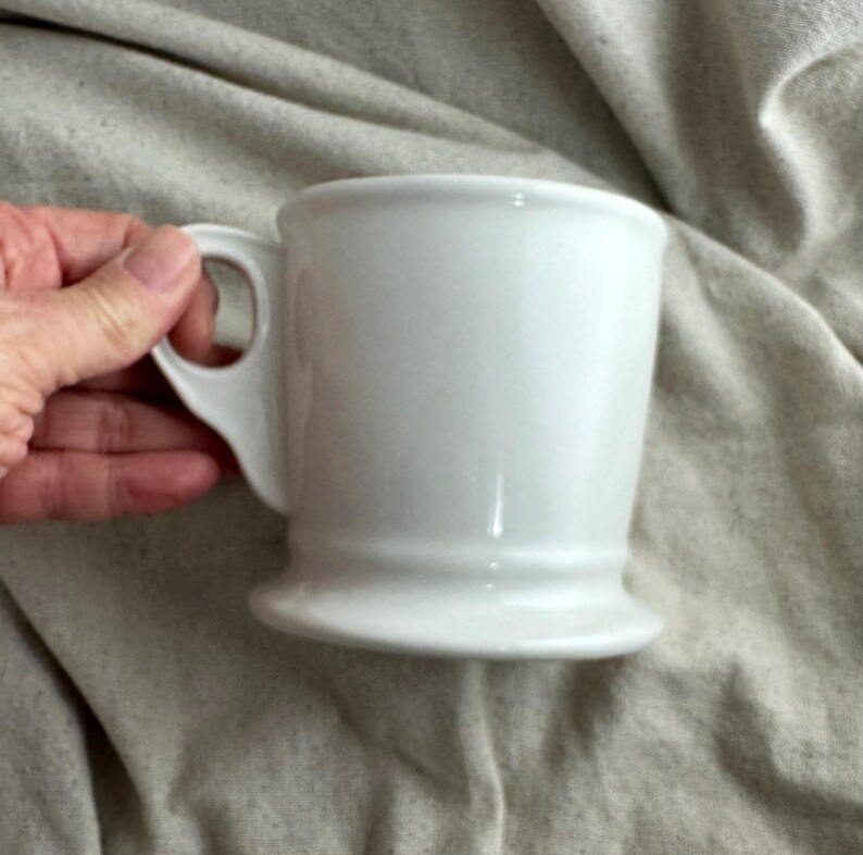 May include: A white ceramic mug with a handle. The mug has a flared shape and a wide base. The handle is a simple loop. The mug is held by a hand against a neutral fabric background.