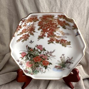 May include: White, hexagonal decorative plate with a gold rim. The plate features a floral and bird design with red, pink, and green accents. The plate is displayed on a brown wooden stand.