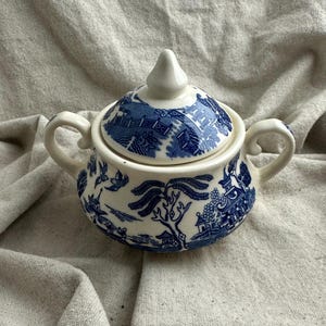 May include: A blue and white ceramic sugar bowl with a lid. The bowl has two handles and a detailed Willow pattern design. The lid has a small, pointed finial. The bowl is approximately 4 inches in diameter.