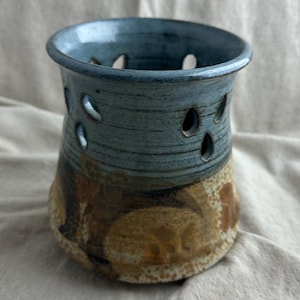 May include: A handmade ceramic candle holder with a blue top and a brown and tan base. The candle holder has several oval-shaped cutouts around the sides. The ceramic piece is likely used for home decor.