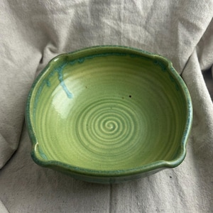 May include: A handmade ceramic bowl in shades of green. The bowl has a spiral design in the center and a scalloped rim. The exterior is a lighter green, while the rim has a darker green glaze with blue accents.