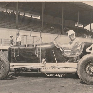 May include: A black and white photo of a vintage race car with the number 4 on the side. The driver is wearing a white helmet and is smiling. The car is on a dirt track and there are spectators in the background.