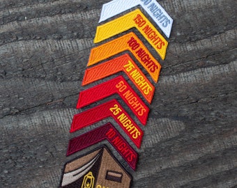 May include: A set of colorful embroidered patches in the shape of arrows, each with a different number of nights printed on them. The patches are arranged in a stack, with the largest patch at the top and the smallest patch at the bottom. The patches are all red, orange, yellow, and white. The bottom patch is a brown tent with a lantern and the word "CAMPER" printed on it.