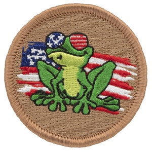 May include: A green frog wearing a red, white, and blue hat sits in front of a distressed American flag. The frog is embroidered on a brown fabric patch.