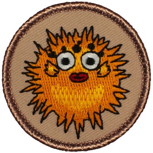 May include: A brown embroidered patch with a cartoon pufferfish with orange and yellow fur, black eyes, and a red mouth. The pufferfish is surrounded by a brown border.