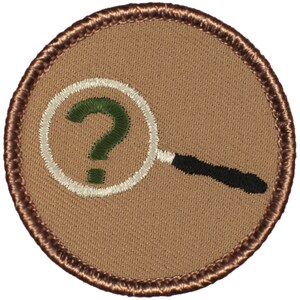 May include: A brown embroidered patch with a white magnifying glass and a green question mark inside the lens. The patch has a brown stitched border.