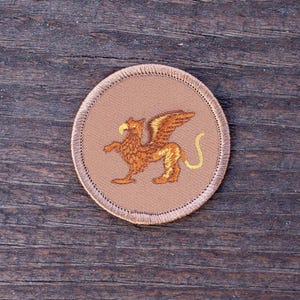 May include: A brown embroidered patch with a gold stitched griffin design. The griffin is a mythical creature with the head and wings of an eagle and the body of a lion.