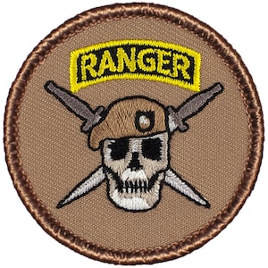 Puede incluir: Un parche circular con fondo marrón y borde cosido marrón. El parche presenta una calavera con boina, cuchillos cruzados y una pancarta amarilla con la palabra "RANGER" en negro.
