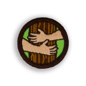 May include: A brown and green embroidered patch with two hands holding a brown tree trunk. The patch is circular and has a brown border.