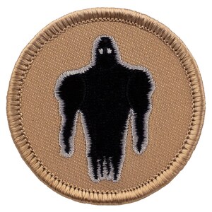 May include: A tan embroidered patch with a black silhouette of a humanoid figure with a white outline. The figure has a large head and small limbs.