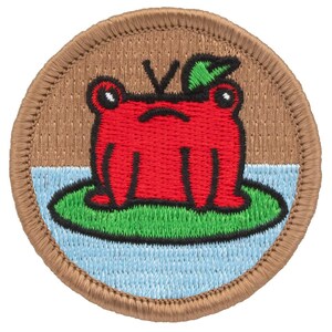 May include: Embroidered patch featuring a red frog on a lily pad. The frog has black eyes and a green leaf on its head. The patch is round with a brown border and a light blue and green background.