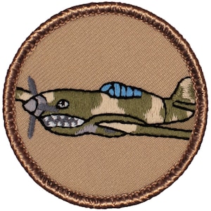 May include: A brown embroidered patch with a green, tan, and blue camouflage airplane on a tan background.