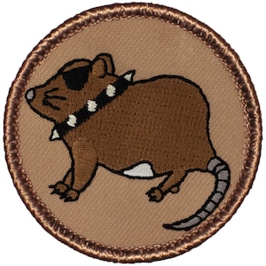 May include: A brown embroidered patch with a cartoon rat wearing a studded collar and sunglasses. The rat is facing left and has a long tail. The patch is circular and has a brown border.