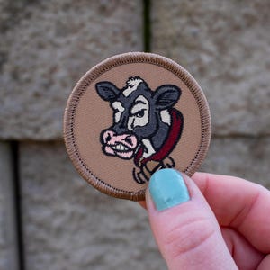 May include: A brown embroidered patch with a black and white cow head design. The cow has a pink nose and a red and white collar.