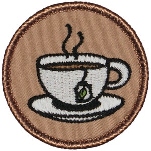 May include: A brown embroidered patch with a white teacup and saucer with a green tea bag. The teacup has steam coming from it.