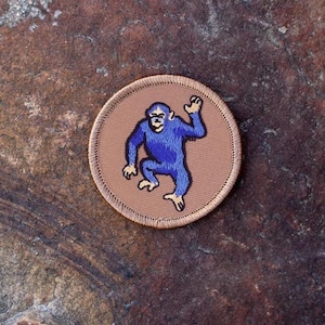 May include: A brown embroidered patch with a blue monkey in a playful pose. The monkey is outlined in black and has a white face.