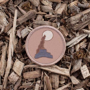 May include: A brown embroidered patch with a wolf howling at the moon. The wolf is silhouetted against a light brown background with a white moon.