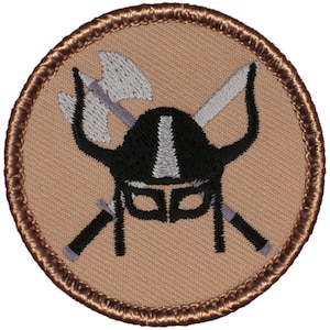 May include: Round patch featuring a black Viking helmet with a white stripe, crossed swords, and axes. The patch has a tan background with a brown stitched border. The design is a stylized emblem.