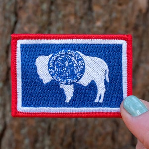 May include: A blue and white embroidered patch with the state seal of Montana, featuring a white bison with a blue circle around it. The patch has a red border.