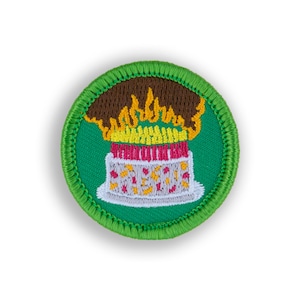 May include: A green embroidered patch with a birthday cake with lit candles and brown frosting. The cake is on a white plate.