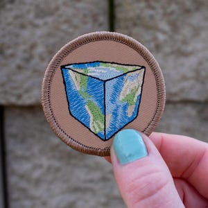 May include: A brown embroidered patch with a blue and green globe inside a cube shape.