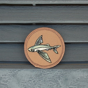 May include: A brown embroidered patch with a flying fish design. The fish is green, blue, and white with brown wings.