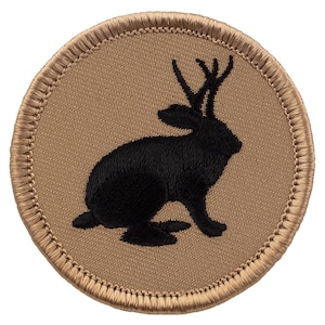May include: A black embroidered patch with a silhouette of a rabbit with antlers on a tan background. The patch has a brown stitched border.