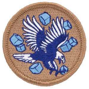 May include: A brown embroidered patch with a blue and white eagle flying with its wings spread. The eagle is surrounded by light blue geometric shapes.