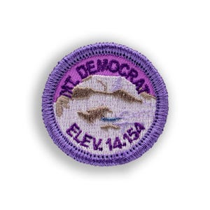 May include: A round, embroidered patch with a purple border and a white center. The center features a mountain scene with the text "MT. DEMOCRAT" and "ELEV 14154" in purple. The patch is likely for a hiking or outdoor activity.
