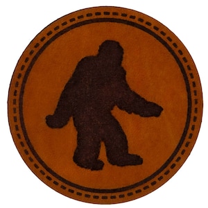 May include: A brown leather coaster with a black silhouette of Bigfoot walking. The coaster has a decorative border of black stitching.
