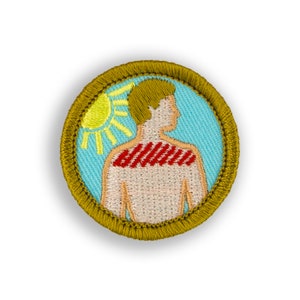 May include: A round, embroidered patch with a gold border. The patch depicts a person with red stripes on their back, standing in front of a yellow sun on a blue background.