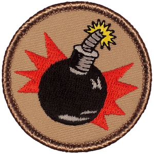 May include: Embroidered patch featuring a black bomb with a lit fuse and a red burst design. The bomb is centered on a tan background, surrounded by a brown border. The patch is circular in shape.