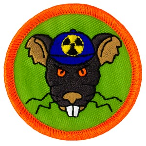 May include: A green and orange embroidered patch featuring a cartoon rat wearing a blue baseball cap with a yellow and black radiation symbol. The rat has orange eyes and white teeth.
