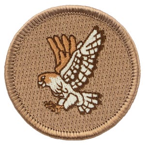 May include: A round, embroidered patch featuring a hawk in flight. The hawk has brown, white, and tan coloring, with its wings spread wide. The patch has a tan background and a brown border.