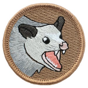 May include: A circular embroidered patch featuring a detailed opossum head in profile against a tan background. The opossum has white fur, black ears, a pink nose, and an open mouth showing sharp white teeth. The patch has a tan stitched border.