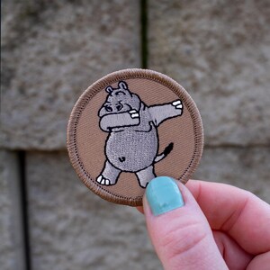 May include: A brown embroidered patch with a gray cartoon hippopotamus doing the dab dance. The patch is circular and has a white outline.