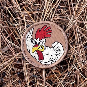 May include: A circular embroidered patch featuring a cartoon rooster flexing its arm. The rooster has a red comb and wattle, a yellow beak, and white feathers. The patch is bordered with brown stitching and set against a background of pine needles.